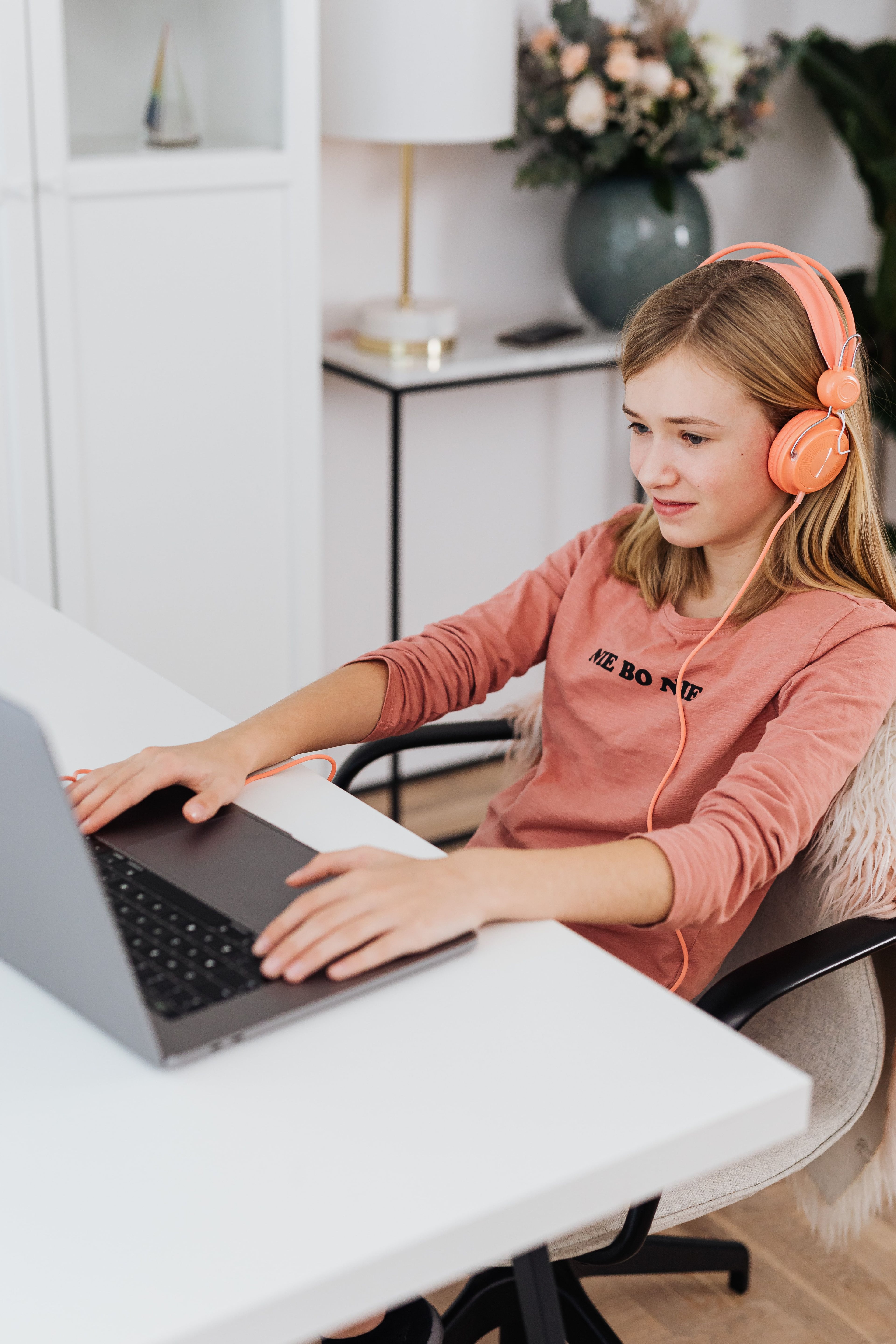 Child with orange headphones using a laptop at a white desk.